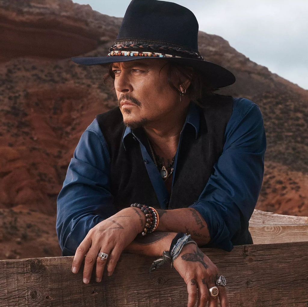Man wearing a black cowboy hat and blue shirt leaning on a wooden fence with desert landscape in the background