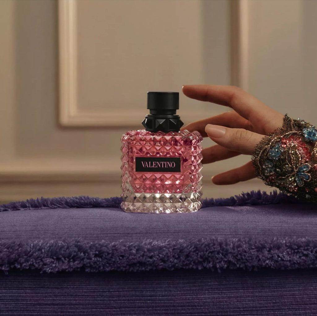 Valentino perfume bottle on a purple surface with a hand reaching towards it.